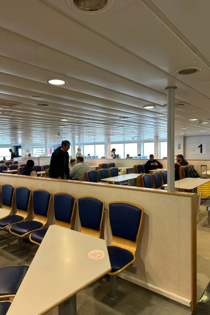 노르웨이 몰데(Molde)로 향하는 페리의 넓고 깨끗한 실내 승객 라운지. 사람들이 편안한 의자에 앉아 휴식을 취하며, 큰 창문 너머로 잔잔한 **피오르드**의 바다 풍경을 바라보고 있다.