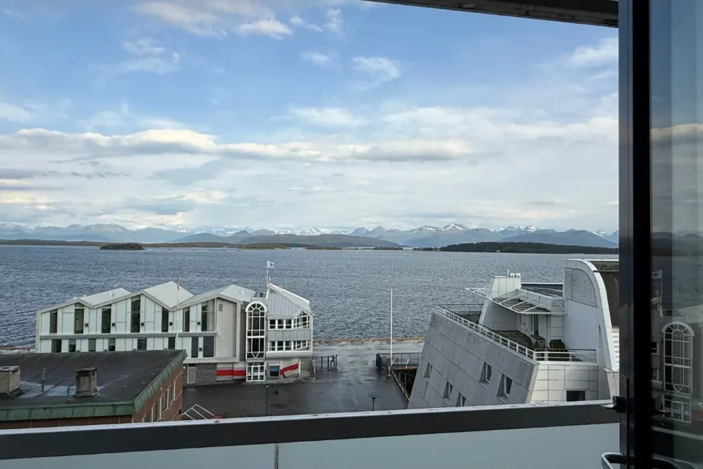 '스칸딕 알렉산드라 몰데' 호텔 객실의 넓은 창문 너머로 그림처럼 펼쳐진 몰데 **피오르드**의 환상적인 오션 뷰. 잔잔한 바다와 건너편의 웅장한 설산이 한눈에 들어온다.